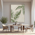 Modern minimalist hand-painted canvas with textured banana leaves in soft green on beige background, displayed in a bright dining room with neutral furniture.
