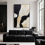 Beige and black abstract wall art adds bold contrast to a black sofa in a modern interior with tall windows and white walls.