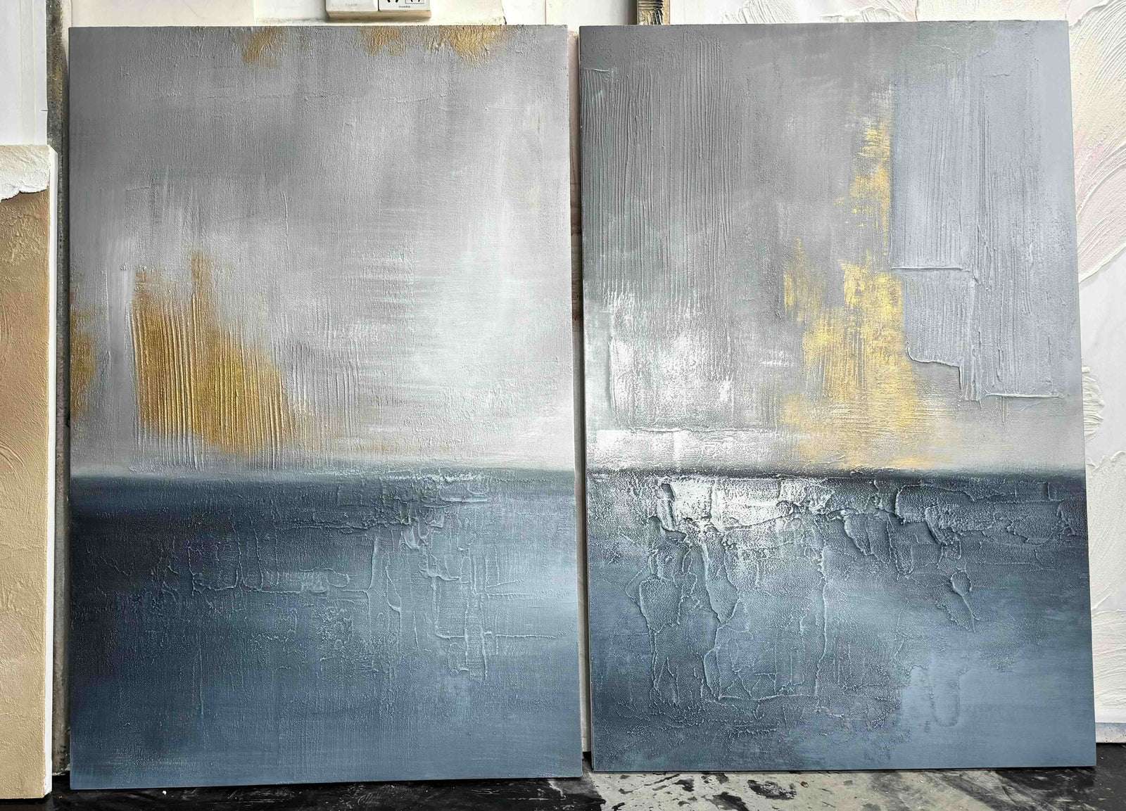 Studio view of two textured abstract seascape canvases in blue gray and gold